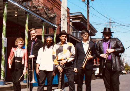 A seven-member band posing on a sunny street with vintage outfits and a drum, guitar, and brass instruments, ready for a performance.