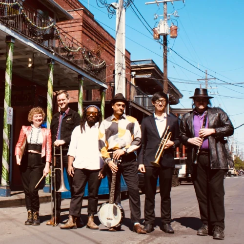A seven-member band posing on a sunny street with vintage outfits and a drum, guitar, and brass instruments, ready for a performance.