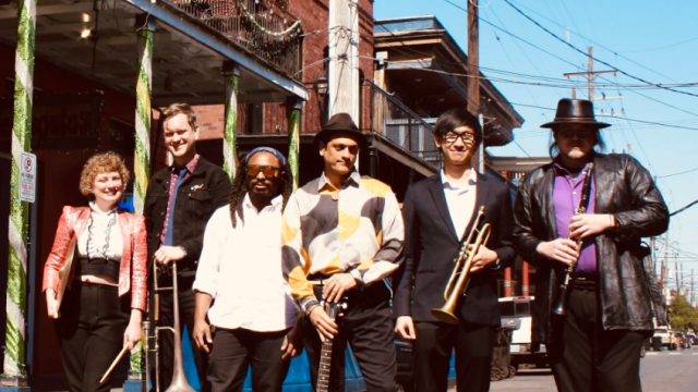 A seven-member band posing on a sunny street with vintage outfits and a drum, guitar, and brass instruments, ready for a performance.