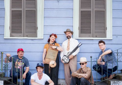 A group of six musicians posing on a porch, one holding a guitar, others with instruments, in front of a blue house with shutters on the windows.