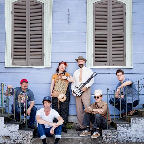 A group of six musicians posing on a porch, one holding a guitar, others with instruments, in front of a blue house with shutters on the windows.
