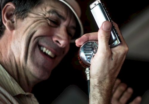 A man wearing a hat is laughing into a vintage-style mic, holding a small handheld device near it as if singing.