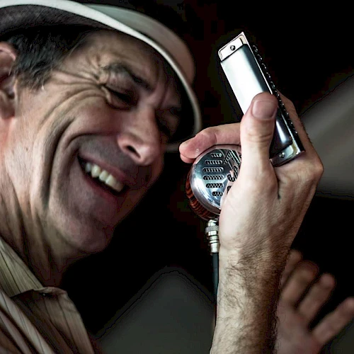 A man wearing a hat is laughing into a vintage-style mic, holding a small handheld device near it as if singing.