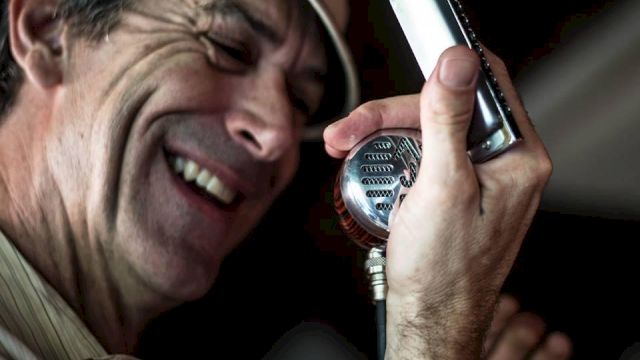 A man wearing a hat is laughing into a vintage-style mic, holding a small handheld device near it as if singing.
