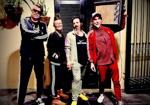 A group of five casually dressed people posing together on a sidewalk, smiling, with one holding a frame or board in the back, sunny vibe.