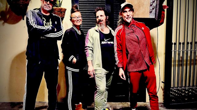 A group of five casually dressed people posing together on a sidewalk, smiling, with one holding a frame or board in the back, sunny vibe.