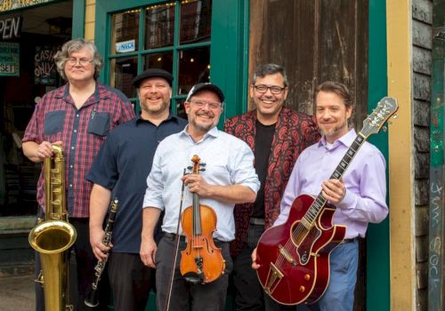 A five-member band posing outside a vintage-style storefront: saxophonist, violinist, guitarist, and two other musicians smiling with instruments, ready to play.