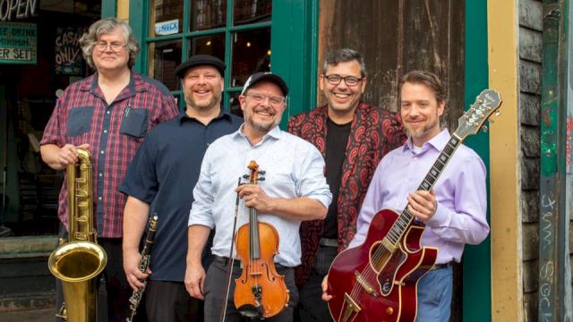 A five-member band posing outside a vintage-style storefront: saxophonist, violinist, guitarist, and two other musicians smiling with instruments, ready to play.