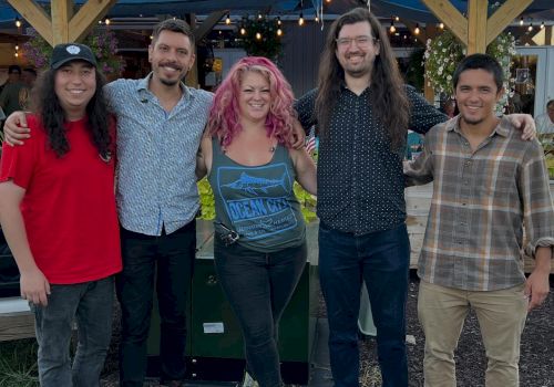 A group of five friends stands outdoors in front of a wooden building, smiling, with casual outfits and relaxed vibes.
