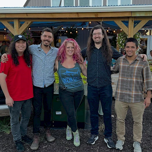 A group of five friends stands outdoors in front of a wooden building, smiling, with casual outfits and relaxed vibes.