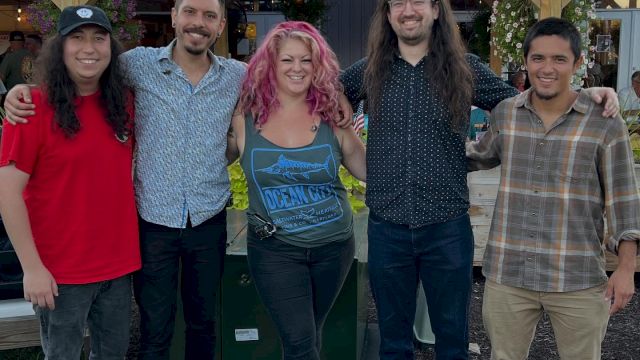 A group of five friends stands outdoors in front of a wooden building, smiling, with casual outfits and relaxed vibes.