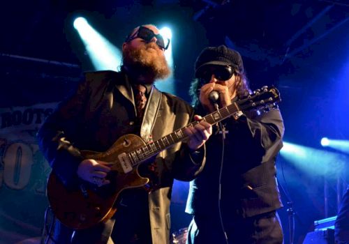 Two guitarists wearing dark suits and sunglasses perform on a dim stage, one singing into a mic while the other plays a guitar.