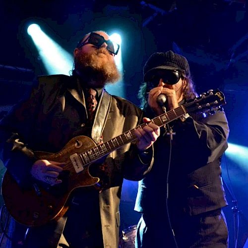 Two guitarists wearing dark suits and sunglasses perform on a dim stage, one singing into a mic while the other plays a guitar.