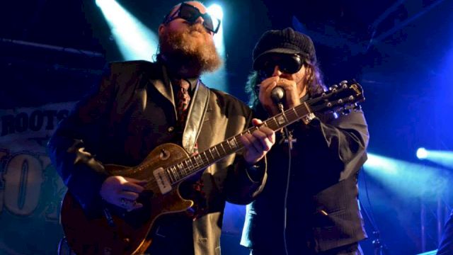 Two guitarists wearing dark suits and sunglasses perform on a dim stage, one singing into a mic while the other plays a guitar.