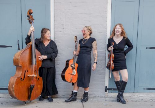 Three women musicians pose with a bass, guitar, and violin in front of blue doors, smiling and ready to perform tonight.