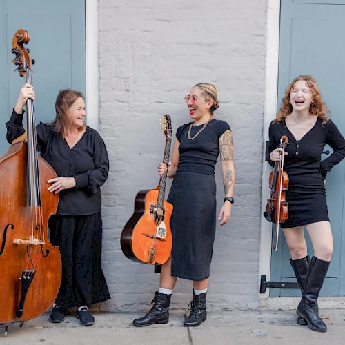 Three women musicians pose with a bass, guitar, and violin in front of blue doors, smiling and ready to perform tonight.