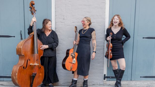 Three women musicians pose with a bass, guitar, and violin in front of blue doors, smiling and ready to perform tonight.