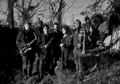 A black-and-white photo of six musicians standing in a yard with trees, holding instruments like saxophone, violin, and trombone, in a moody outdoor scene.