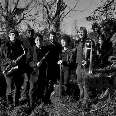 A black-and-white photo of six musicians standing in a yard with trees, holding instruments like saxophone, violin, and trombone, in a moody outdoor scene.