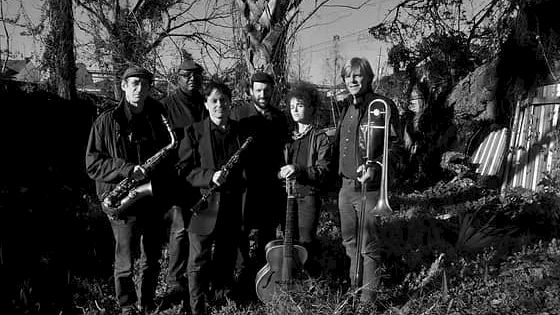 A black-and-white photo of six musicians standing in a yard with trees, holding instruments like saxophone, violin, and trombone, in a moody outdoor scene.