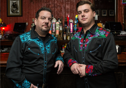 Two men wearing matching black western shirts with blue and red embroidery stand behind a bar, bottles and taps visible in the background.