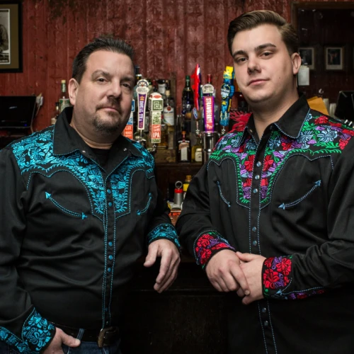 Two men wearing matching black western shirts with blue and red embroidery stand behind a bar, bottles and taps visible in the background.