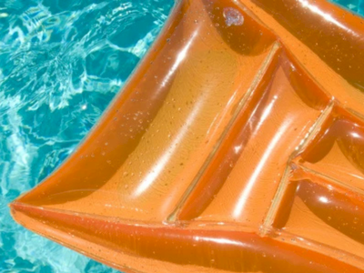 An orange inflatable float in clear blue water, with sunlight reflections on the surface, showing curved seams and a bright pool edge.