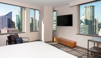 The image shows a modern hotel room with large windows offering a city view, a flat-screen TV, a chair, and a glimpse of a bed.