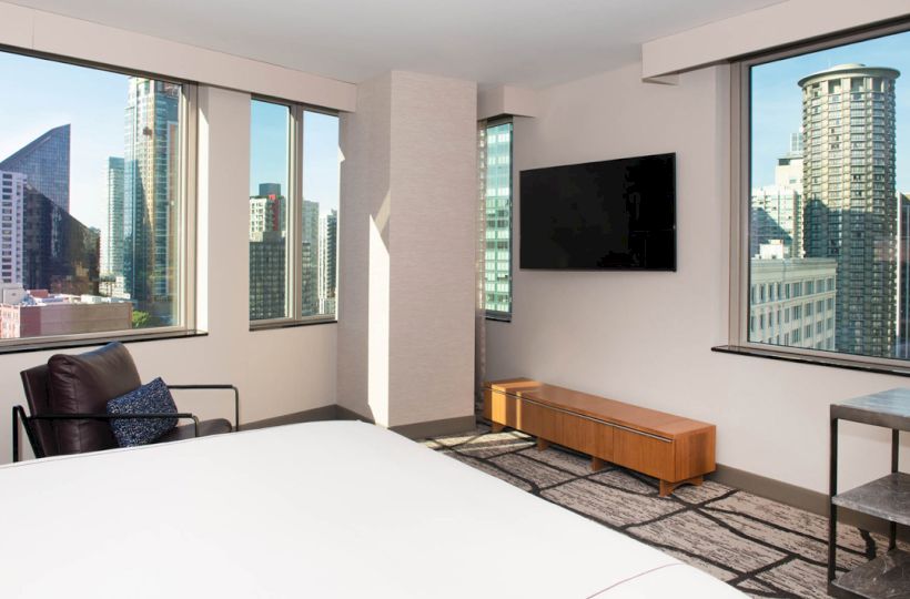 The image shows a modern hotel room with large windows offering a city view, a flat-screen TV, a chair, and a glimpse of a bed.