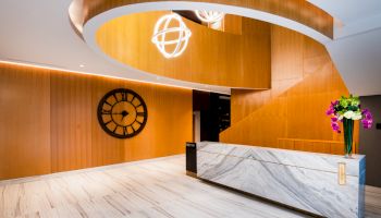 A modern lobby with a curved wooden design, large clock, and sleek marble reception desk adorned with purple flowers.