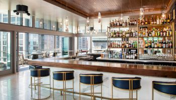 The image shows an elegant bar with stools, a variety of bottles on shelves, and large windows offering a cityscape view.