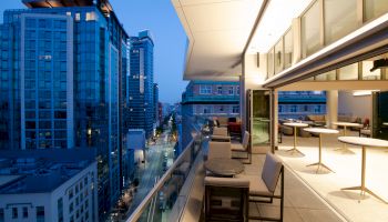 A modern balcony view showing city buildings at dusk with seating and tables, overlooking a street lined with illuminated skyscrapers.