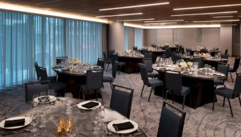 A banquet hall with round tables set elegantly, featuring black chairs and tableware, under soft lighting and surrounded by blue curtains.