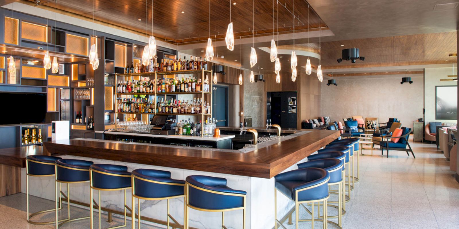 A modern bar with a central counter, stylish chairs, hanging lights, and a fully stocked display, surrounded by a chic lounge area.