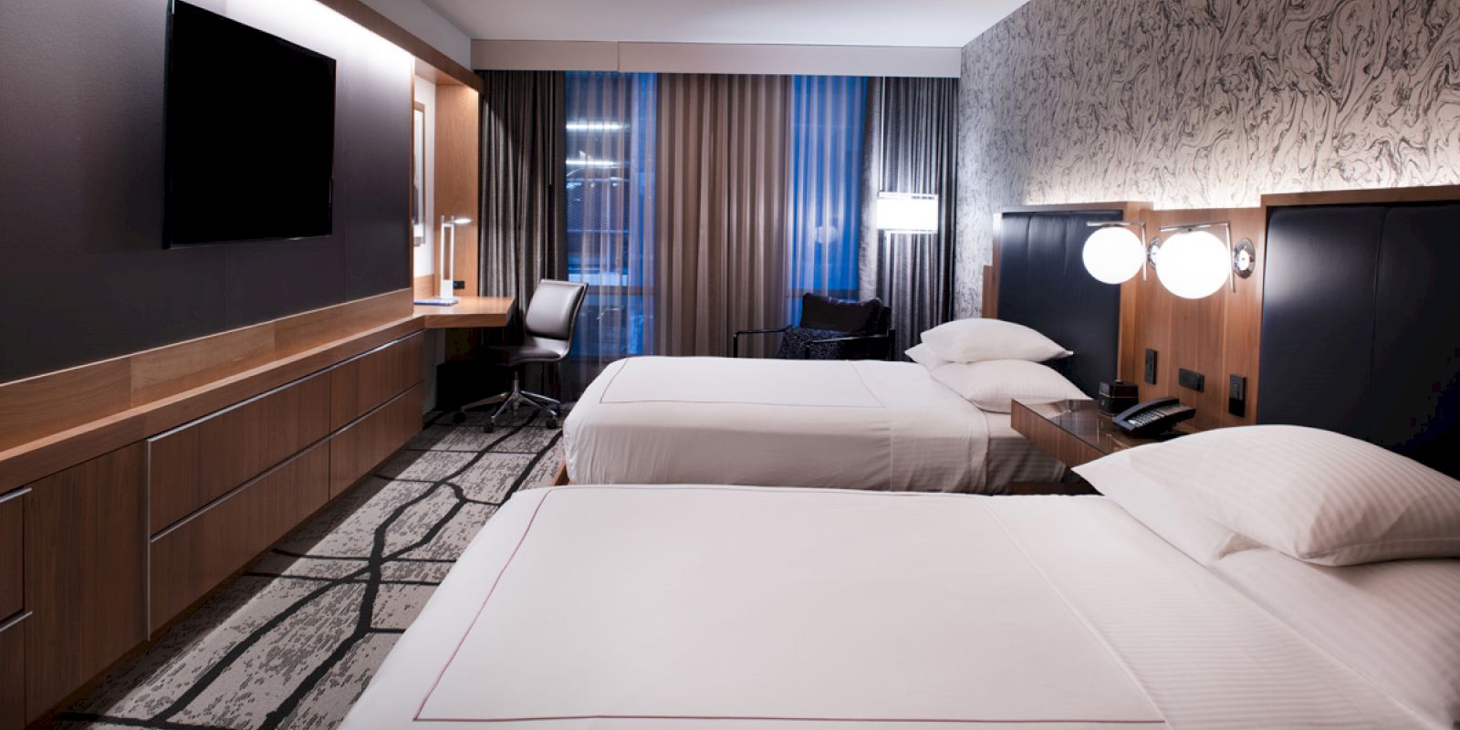 The image shows a modern hotel room with two beds, a TV, desk, and seating area, featuring stylish decor and warm lighting.