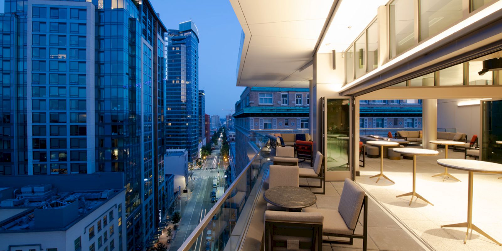The image shows a modern balcony overlooking a city street, with chairs and tables, and tall buildings illuminated at dusk.