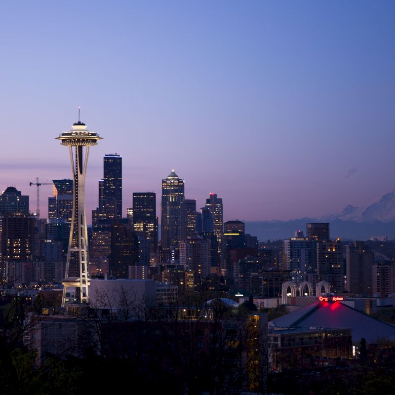 The image shows the Seattle skyline at dusk, featuring the Space Needle and Mount Rainier in the background, under a clear sky.