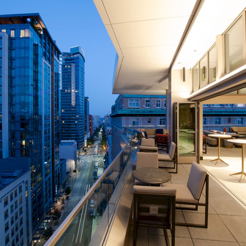A modern rooftop terrace with seating overlooks a cityscape of tall buildings and a street below at twilight.