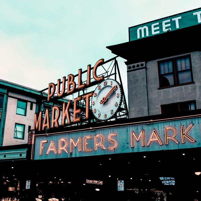 The image shows a famous market entrance with neon signs reading "Public Market" and "Farmers Market," featuring a clock above.