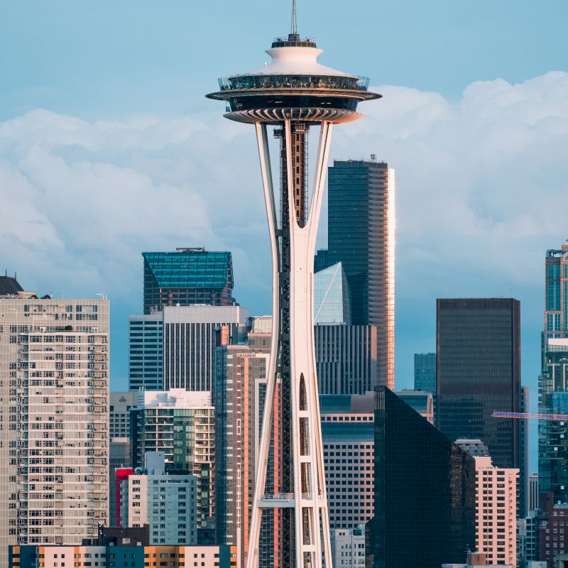 The image shows a city skyline with a prominent structure resembling the Space Needle, surrounded by modern buildings under a cloudy sky.