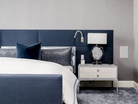 A modern bedroom with a navy bed, white and navy bedding, a bedside table, lamp, and decorative stone piece, all on a gray carpet.