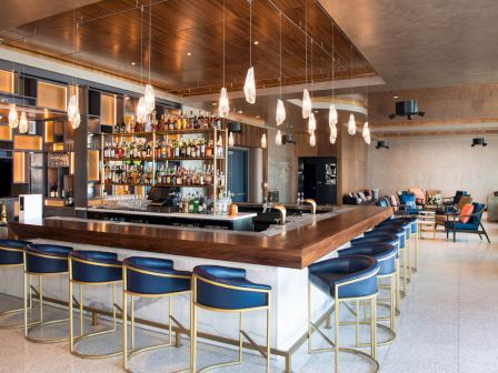 A modern bar with blue stools, overhead pendant lights, and a well-stocked back shelf is surrounded by contemporary seating areas.