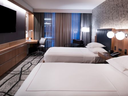 The image shows a modern hotel room with two beds, a TV, a desk and chair, and decorative lighting.