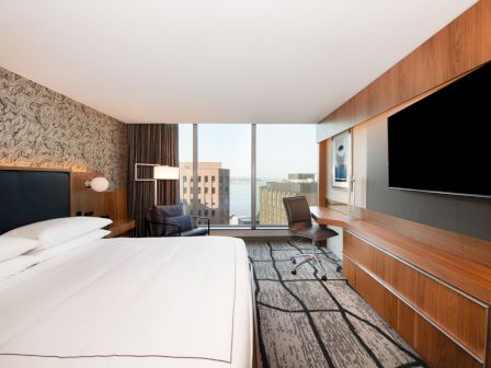 A modern hotel room with a large bed, desk, chair, and television on the wall, overlooking a cityscape through large windows.