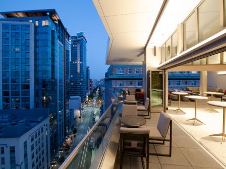 A modern balcony overlooking a cityscape at dusk, with tables and chairs, glass railings, and tall buildings lining the street below.