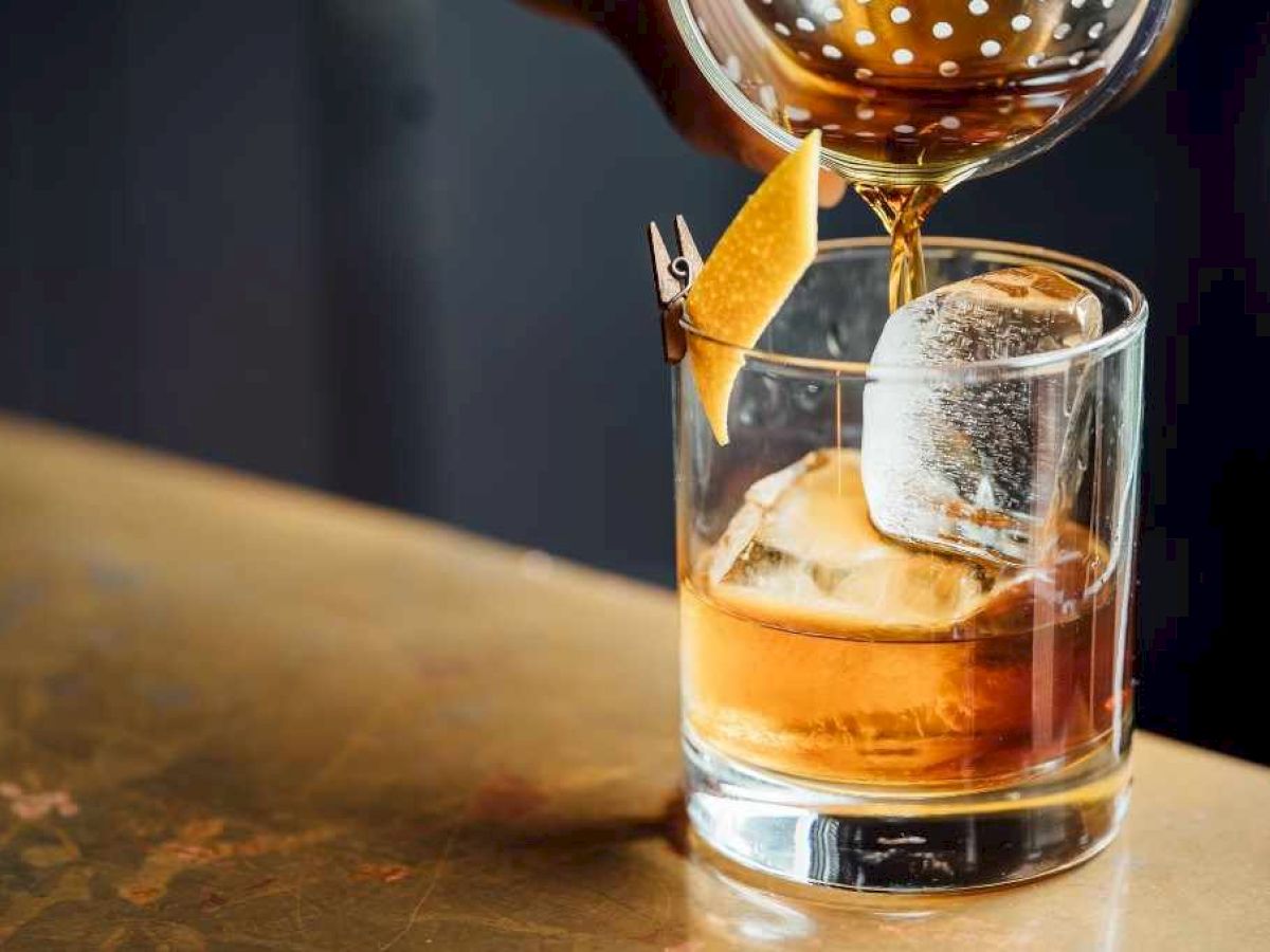 A drink is being poured into a glass with ice and a lemon twist garnish, placed on a table.
