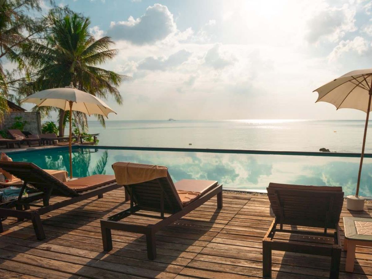 A tranquil beach setting with lounge chairs and umbrellas on a wooden deck overlooking a calm sea with palm trees and a bright sky.