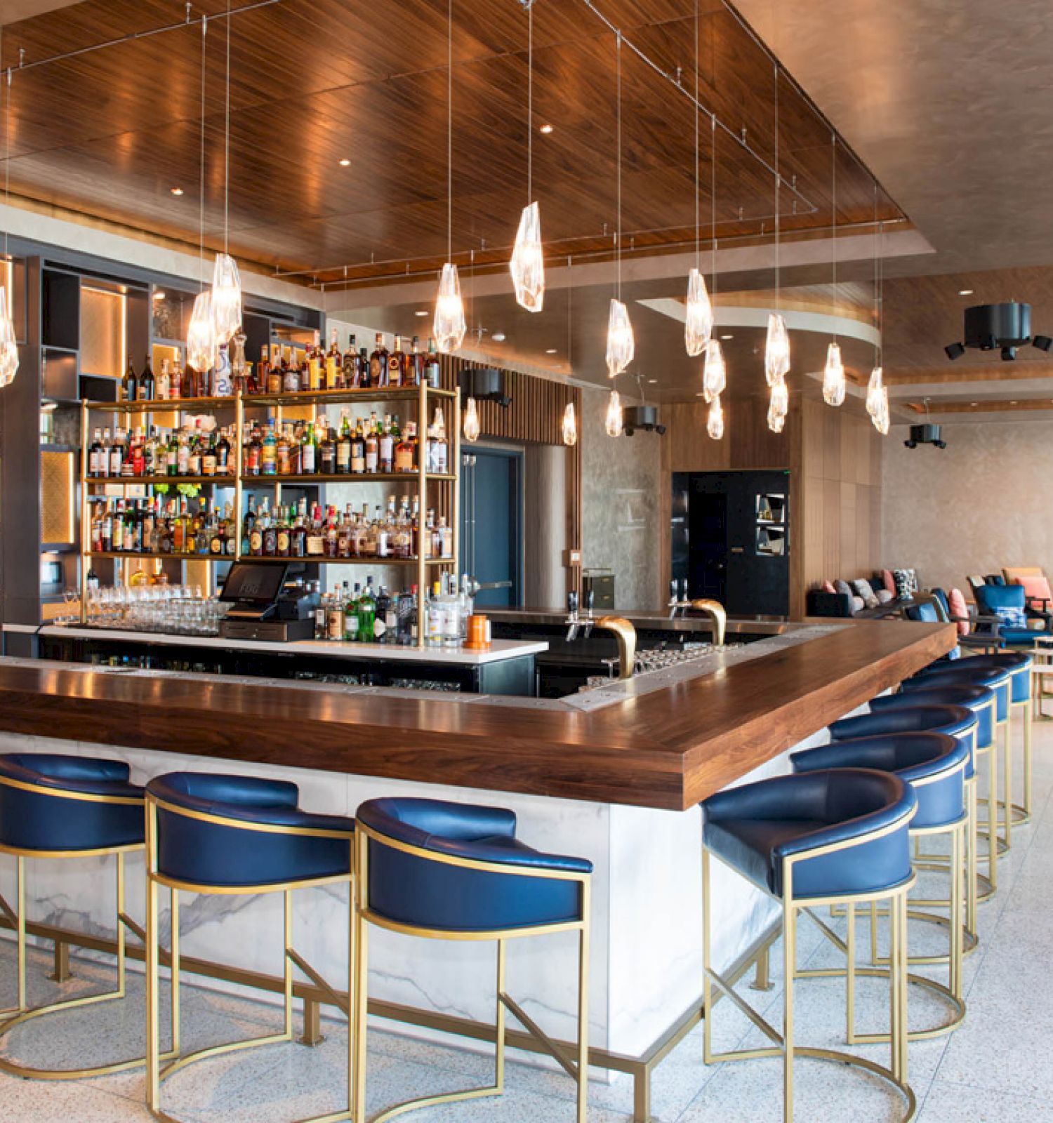A modern bar with hanging lights, a fully stocked shelf, and blue seating around a wooden counter, in a spacious lounge setting.
