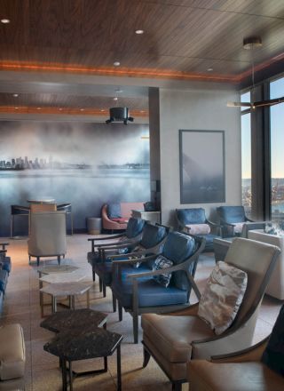 The image shows a modern lounge with plush seating, a wall mural, and large windows offering a view of a waterfront and a ferris wheel.