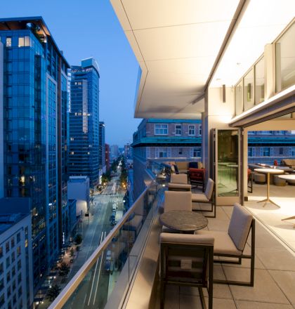 A cityscape showing buildings and a modern outdoor terrace with seating, tables, and a view of the street below, during twilight.